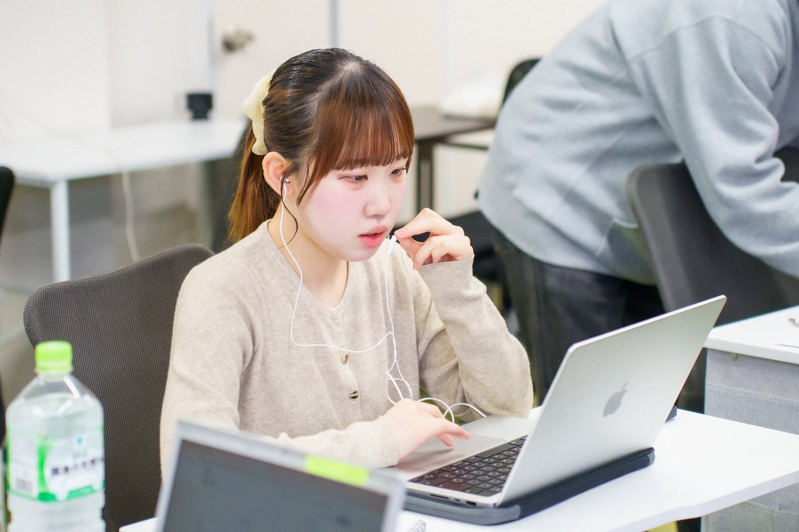 ベージュのカーディガンの女性が、イヤホンのマイクに手を添えて、ノートPCに向かって話している