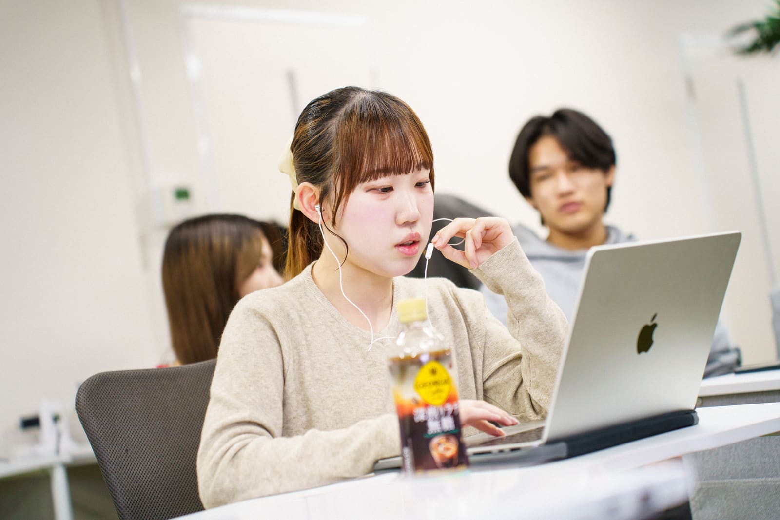 ベージュのカーディガンの女性が、イヤホンのマイクに手を添えて、ノートPCに向かって話している。後ろに男性が座っている