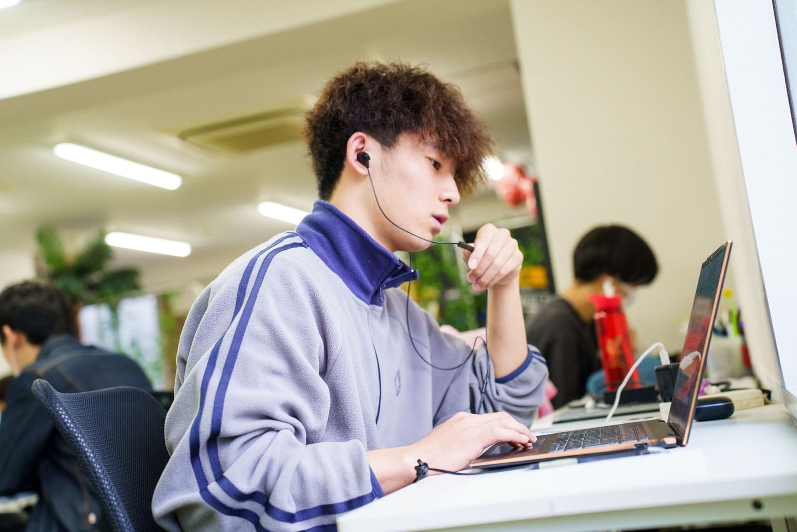 くせ毛の男性が、イヤホンのマイクを口元に寄せて話しながら、ノートPCを操作している横顔