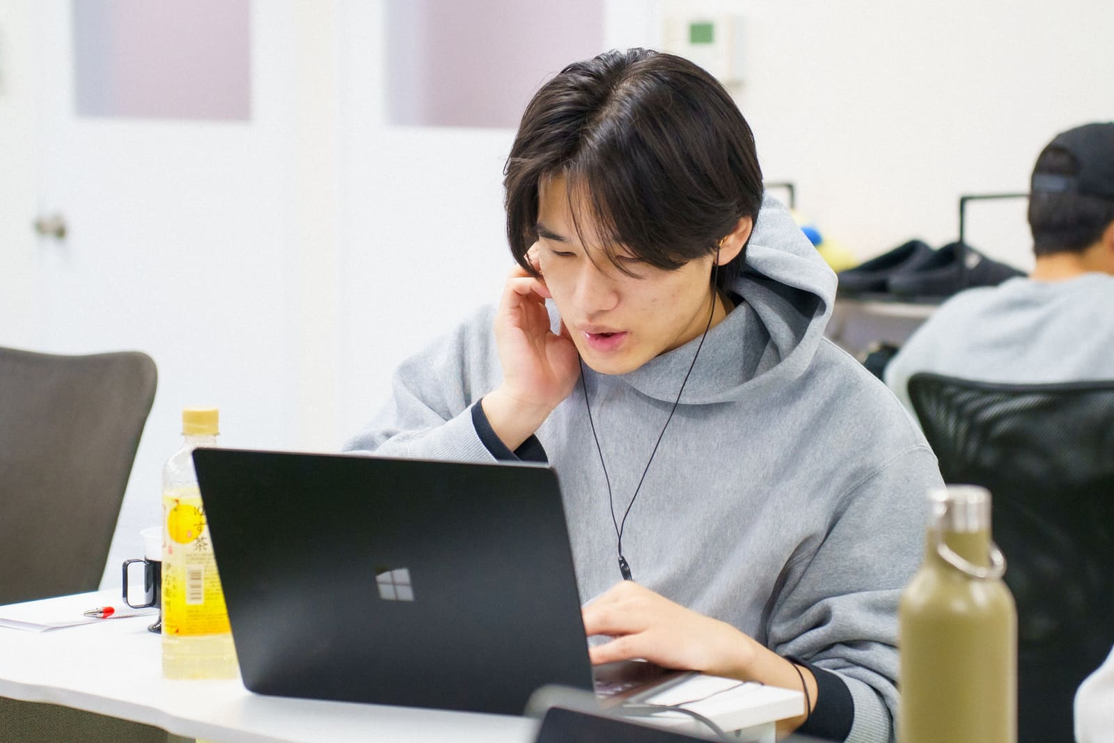 グレーのパーカーの男性が、片手を耳に当て、イヤホンをつけて話しながら、ノートPCを操作している