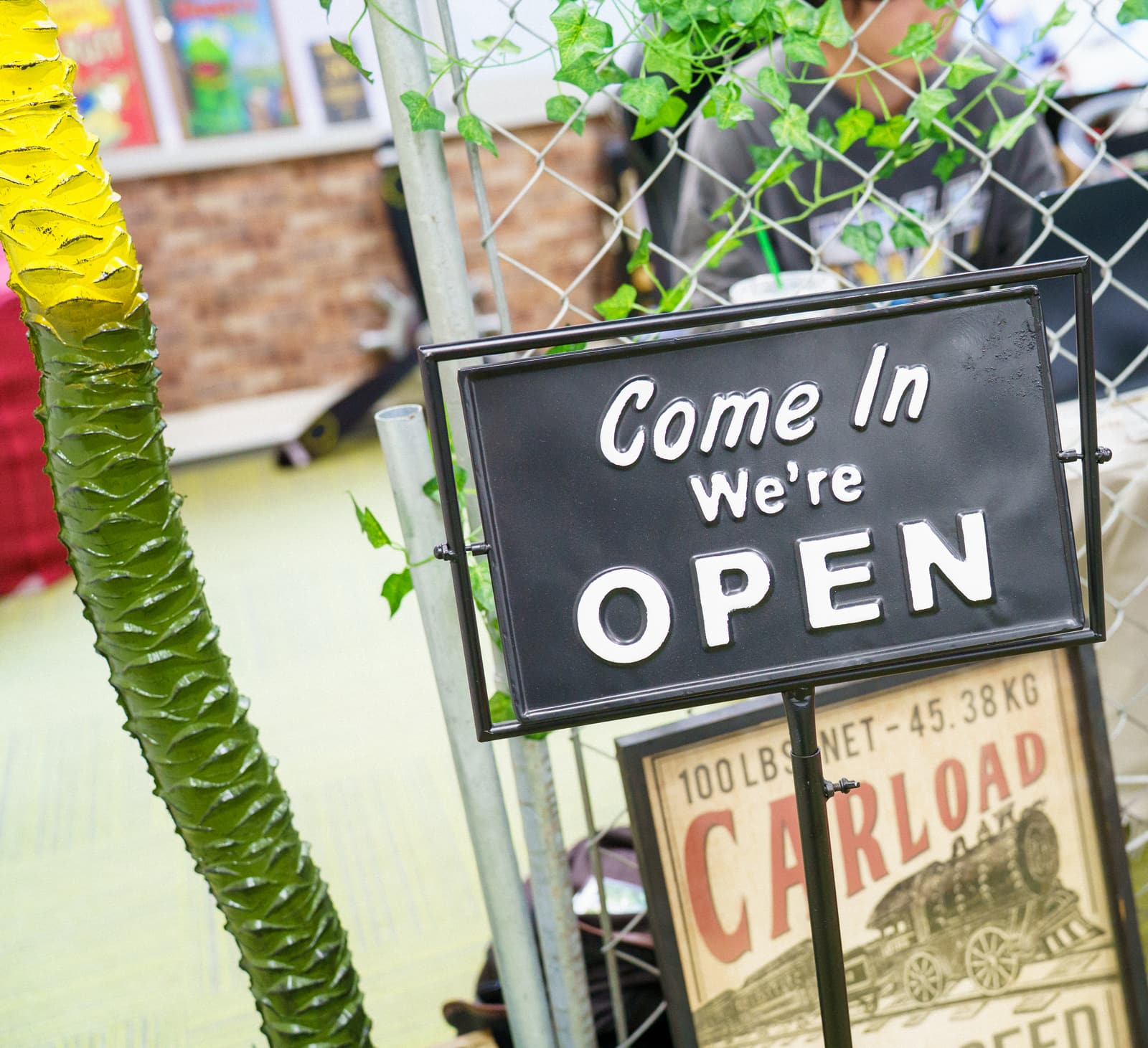 「Come In We're OPEN」と書かれた黒い札。ヤシの木の飾りと、つる草のフェンスの前に置かれている