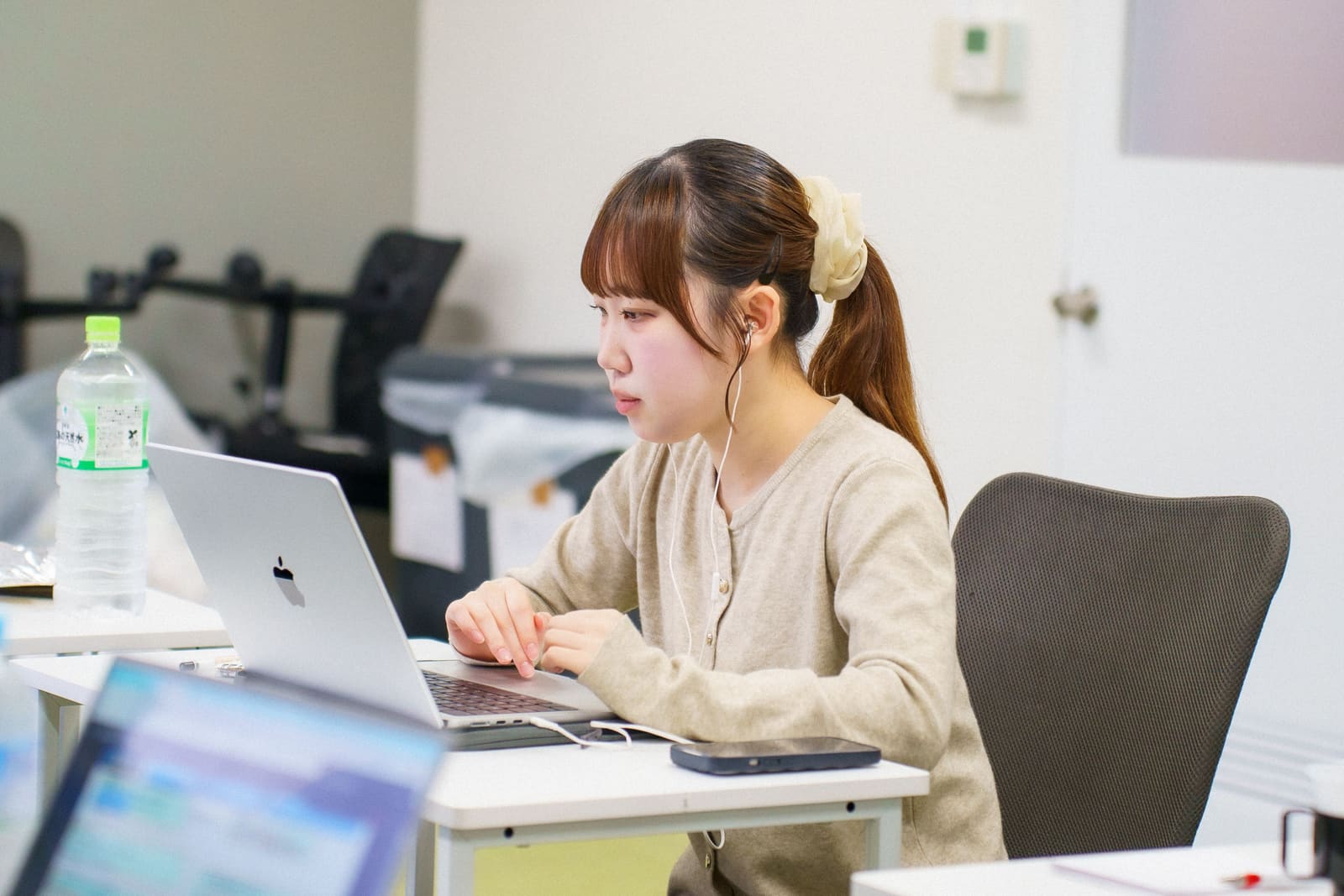 ベージュのカーディガンで髪を結んだ女性が、イヤホンをつけて、ノートPCの画面を見ている横顔