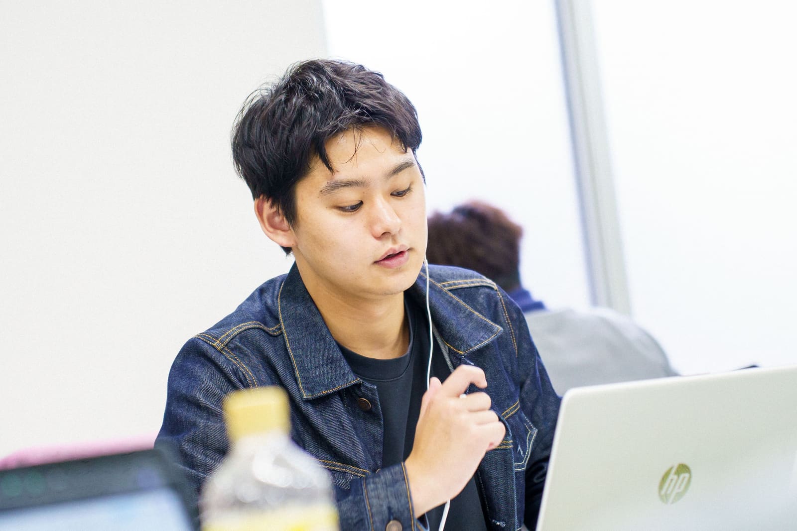 デニムジャケットの男性が、イヤホンをつけて、うつむいて話している。手前にノートPC