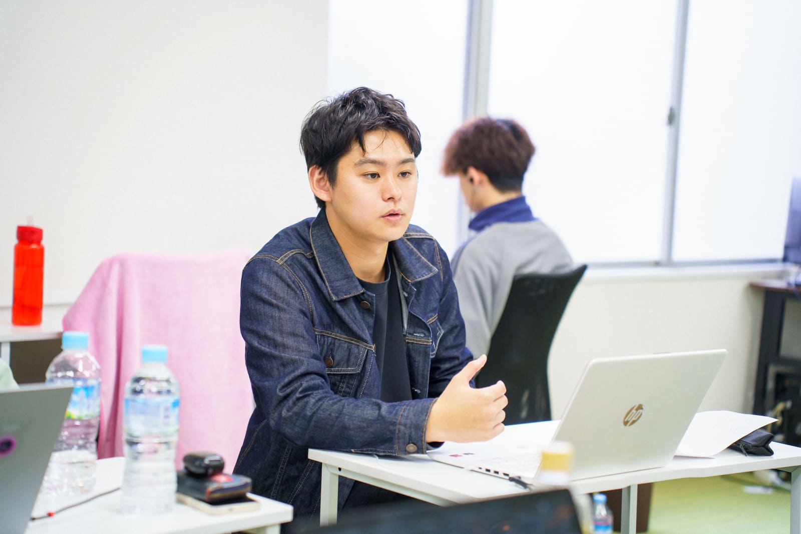デニムジャケットの男性が、ノートPCの前で、手を動かしながら話している。奥に別の男性の背中