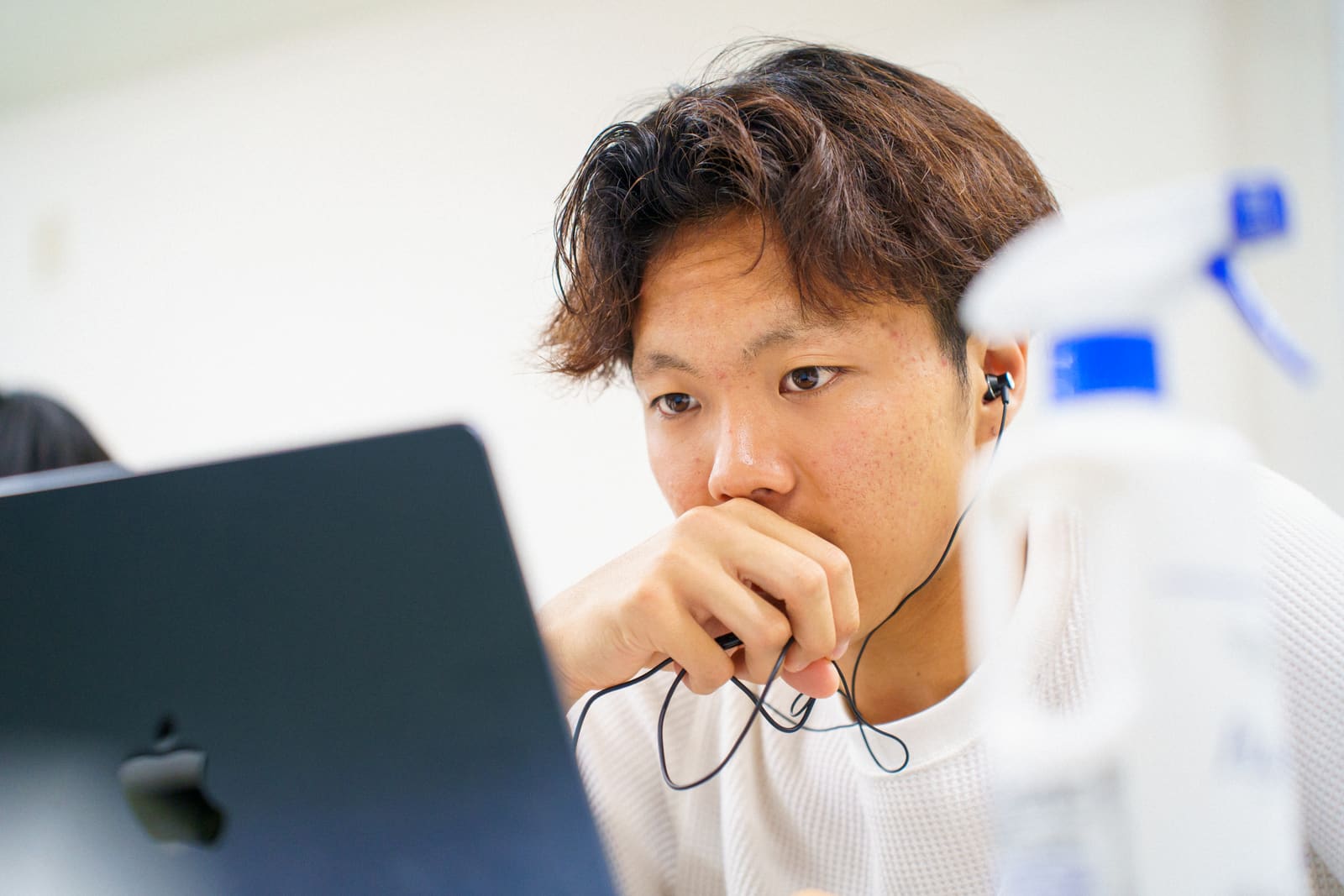 白いニットの男性が、イヤホンのコードを握った手を口元に当てて、ノートPCの画面を見つめている顔のアップ