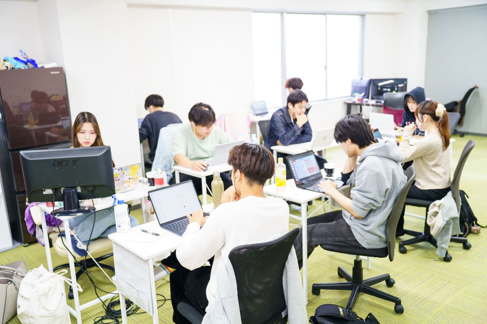 窓から光が入る広いオフィスで、メンバーたちがノートPCに向かう様子を、少し高い位置から見た全景