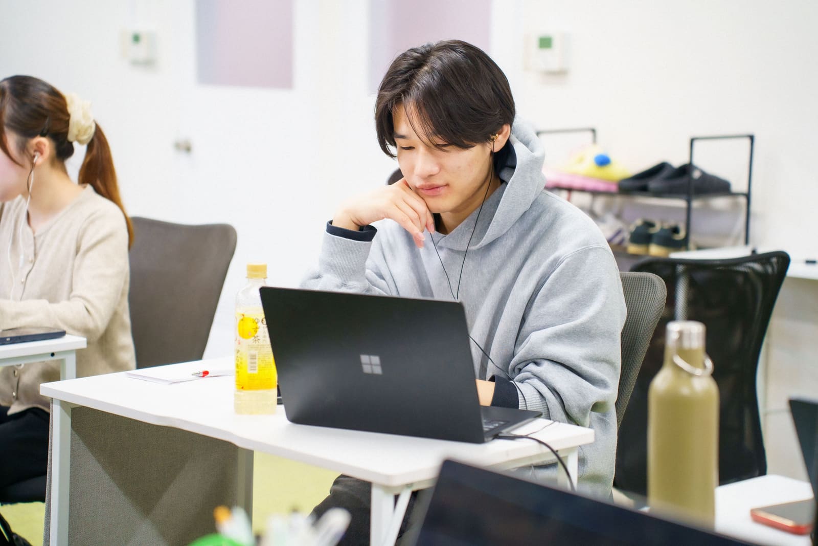 グレーのパーカーの男性が、頬杖をついて、ノートPCの画面を見ている。奥にもう1人