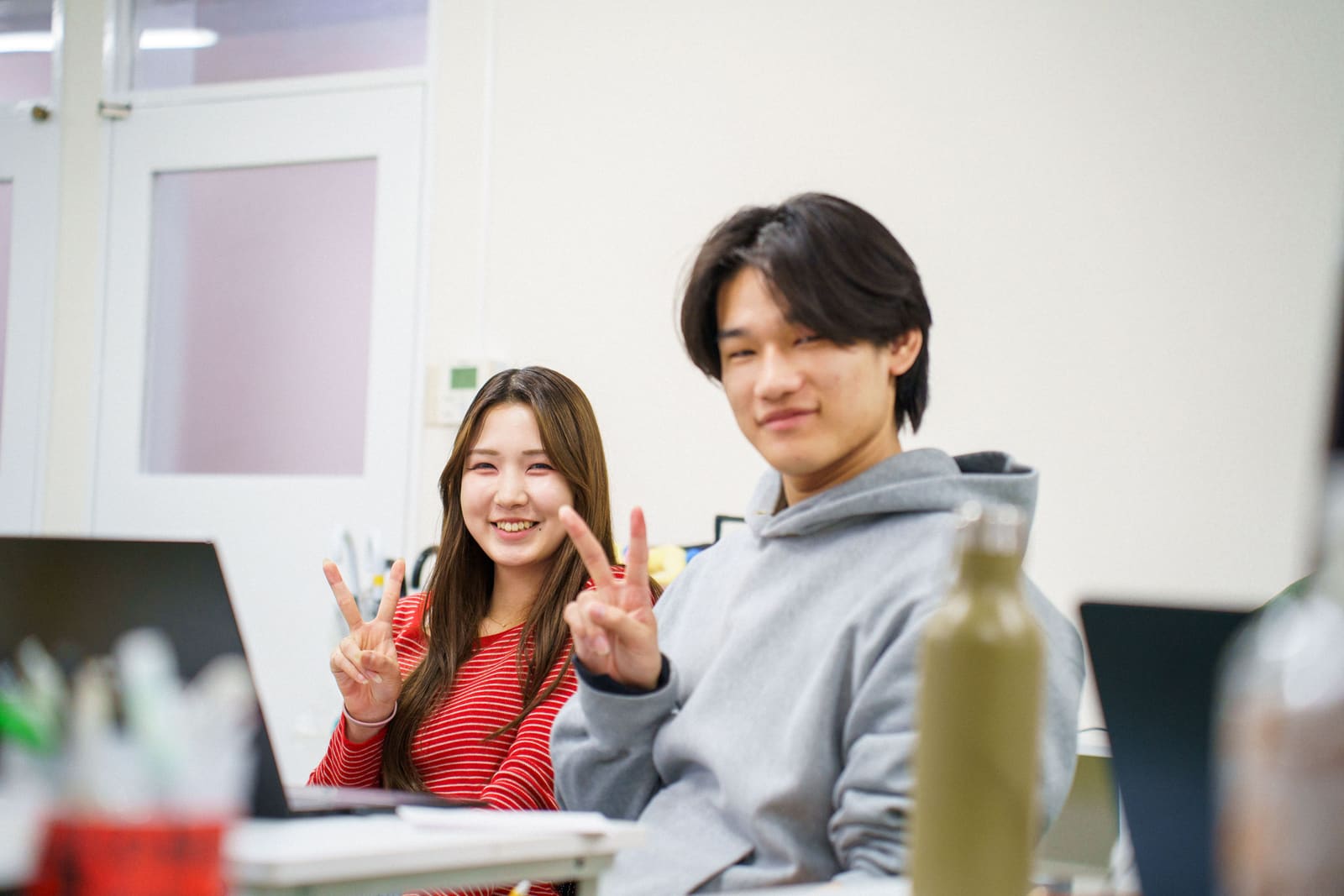 赤いボーダーの服の女性とグレーのパーカーの男性が、並んでこちらを見て、笑顔でピースをしている