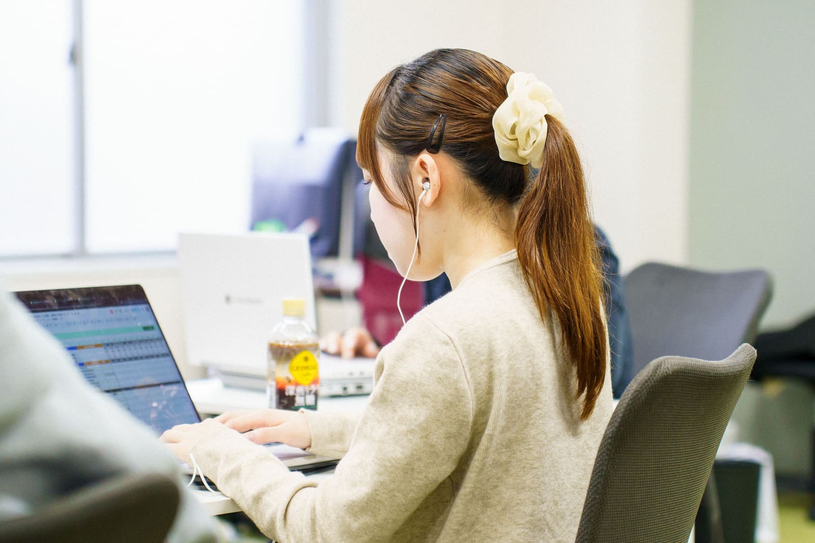 シュシュでポニーテールにしたベージュのニットの女性が、イヤホンをつけて、ノートPCに入力している。斜め後ろから見た姿