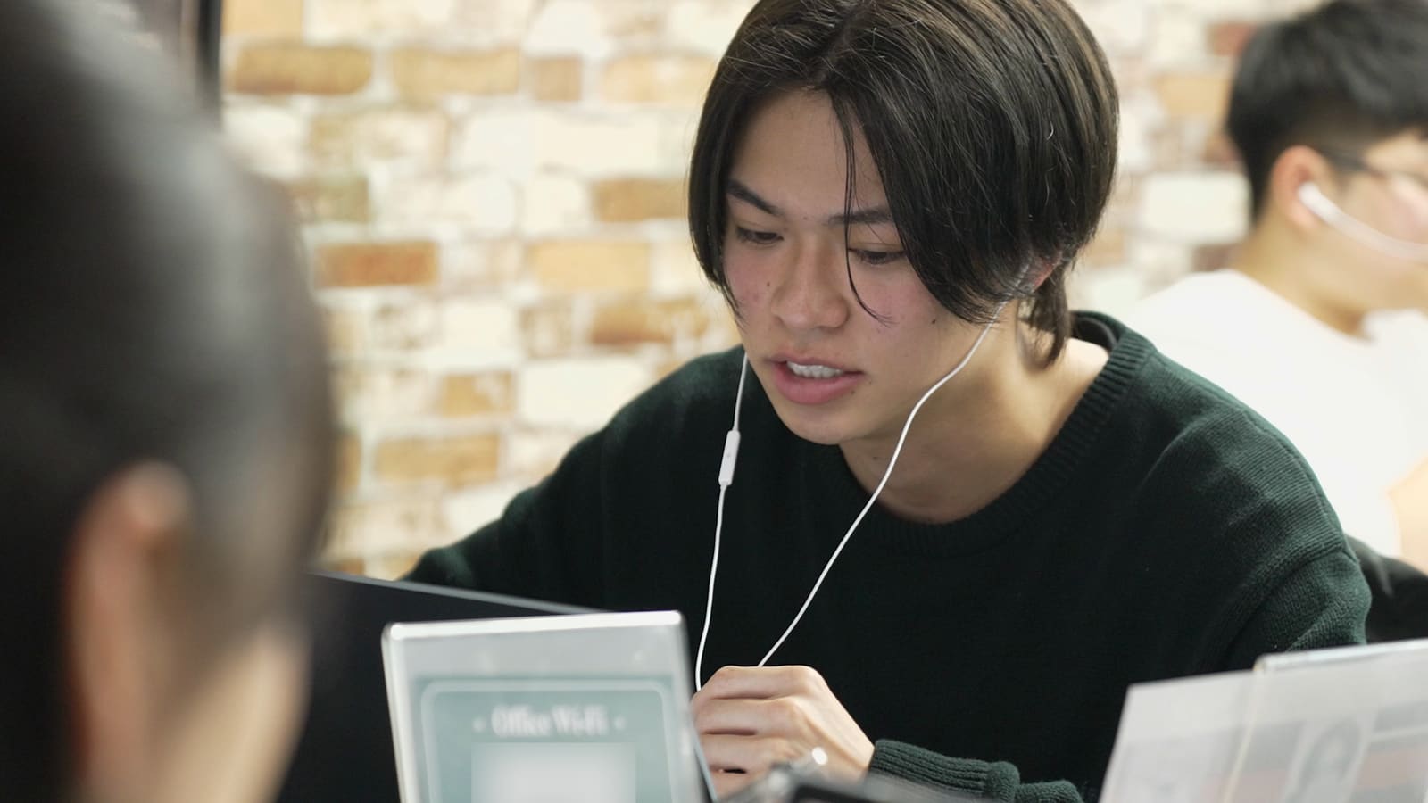深緑のセーターの男性が、イヤホンをつけて、うつむいて話している。レンガの壁の前