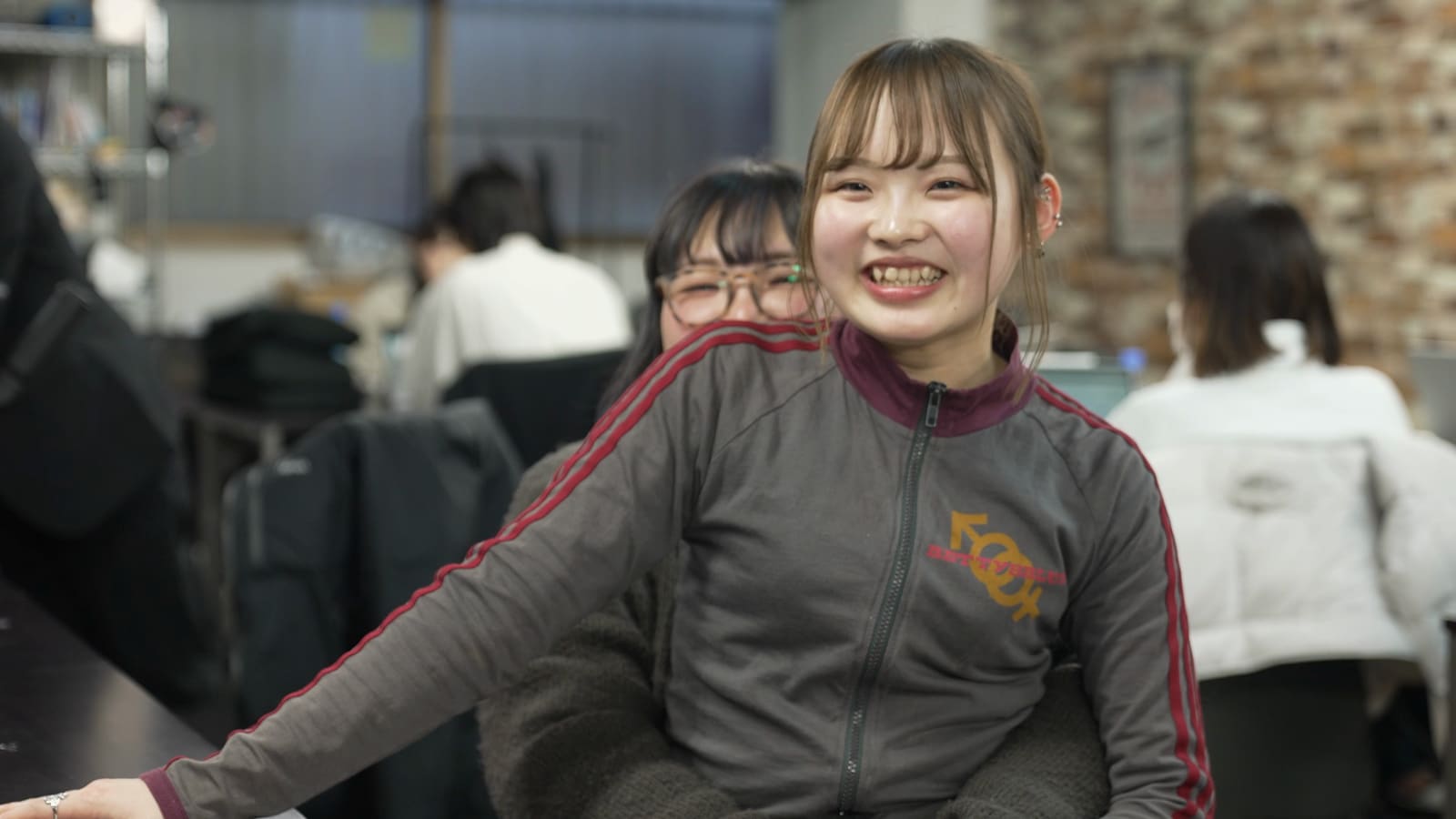赤い線の入ったジャージの女性が、机に手をついて、歯を見せて笑っている。後ろに眼鏡の女性
