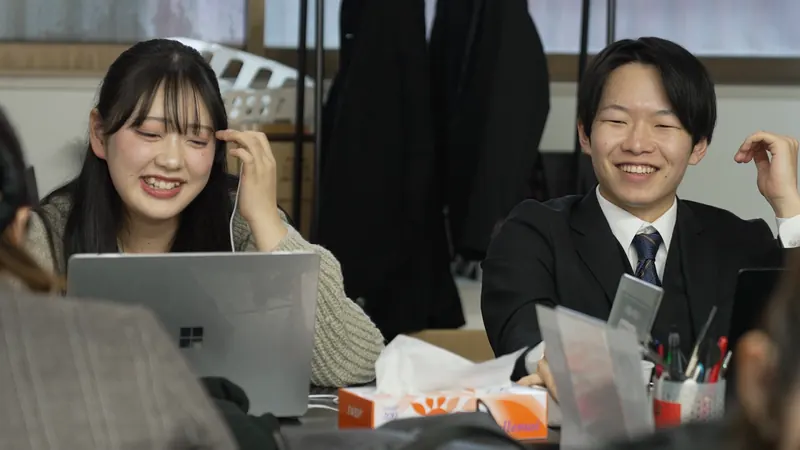 イヤホンのコードを指で持つ女性と、スーツの男性が、ノートPCを前に笑っている