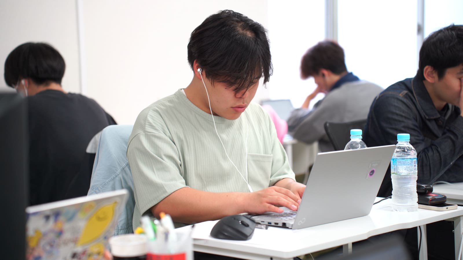 ミント色のTシャツの男性が、イヤホンをつけて、ノートPCに入力している。奥にも作業する人がいる