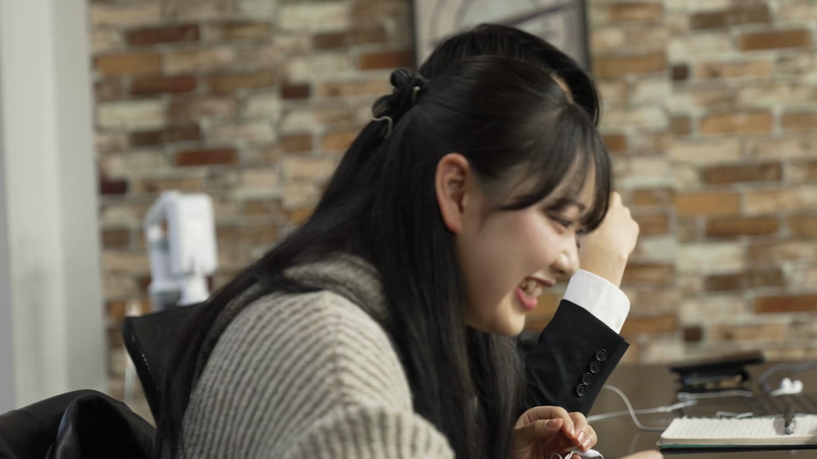 長い黒髪の女性が、ノートPCの前で、口を開けて笑っている。隣にスーツの袖が見える