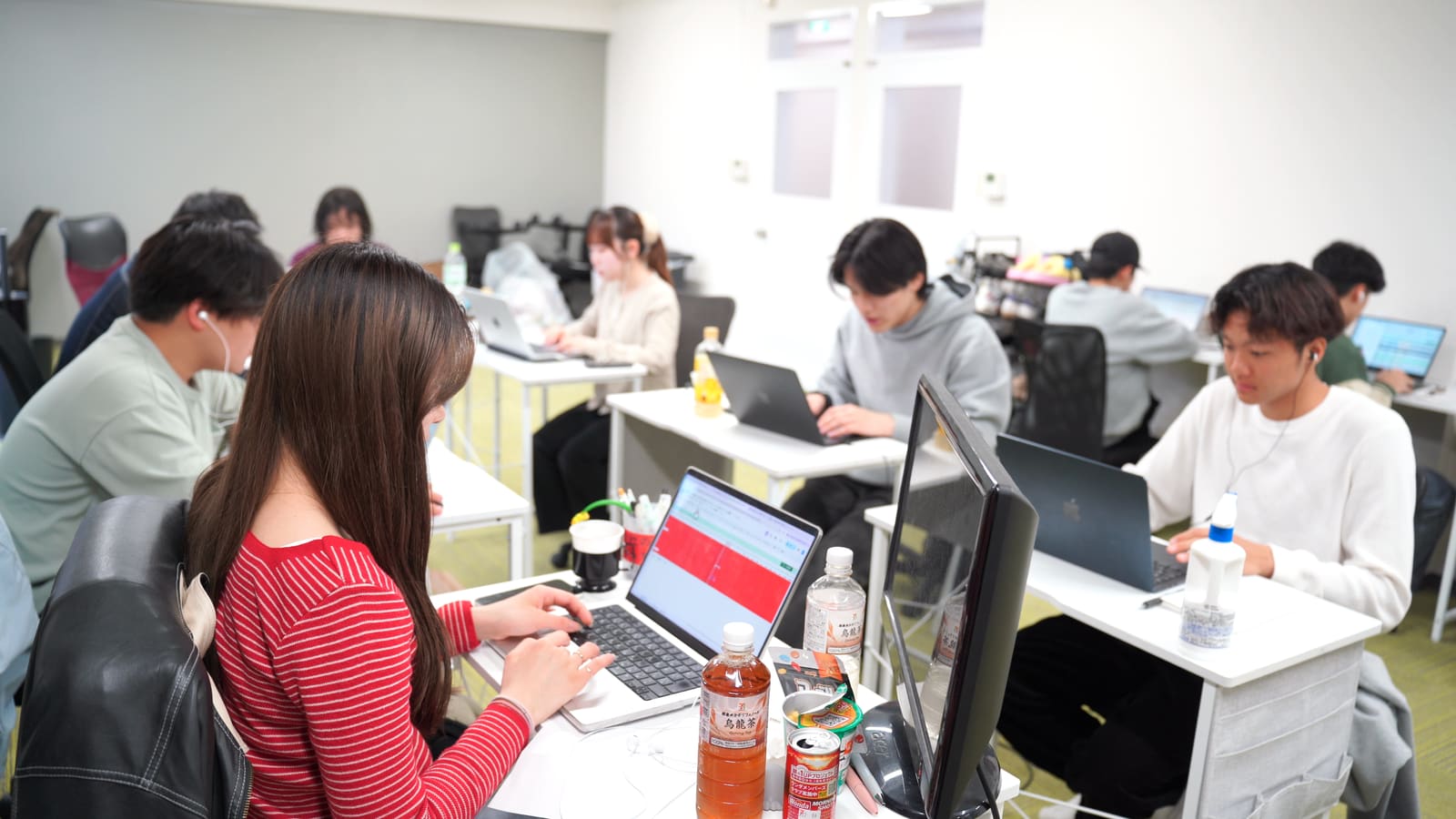 赤いボーダーの長い髪の女性が、ノートPCに入力している。奥の机でも、メンバーたちが作業している