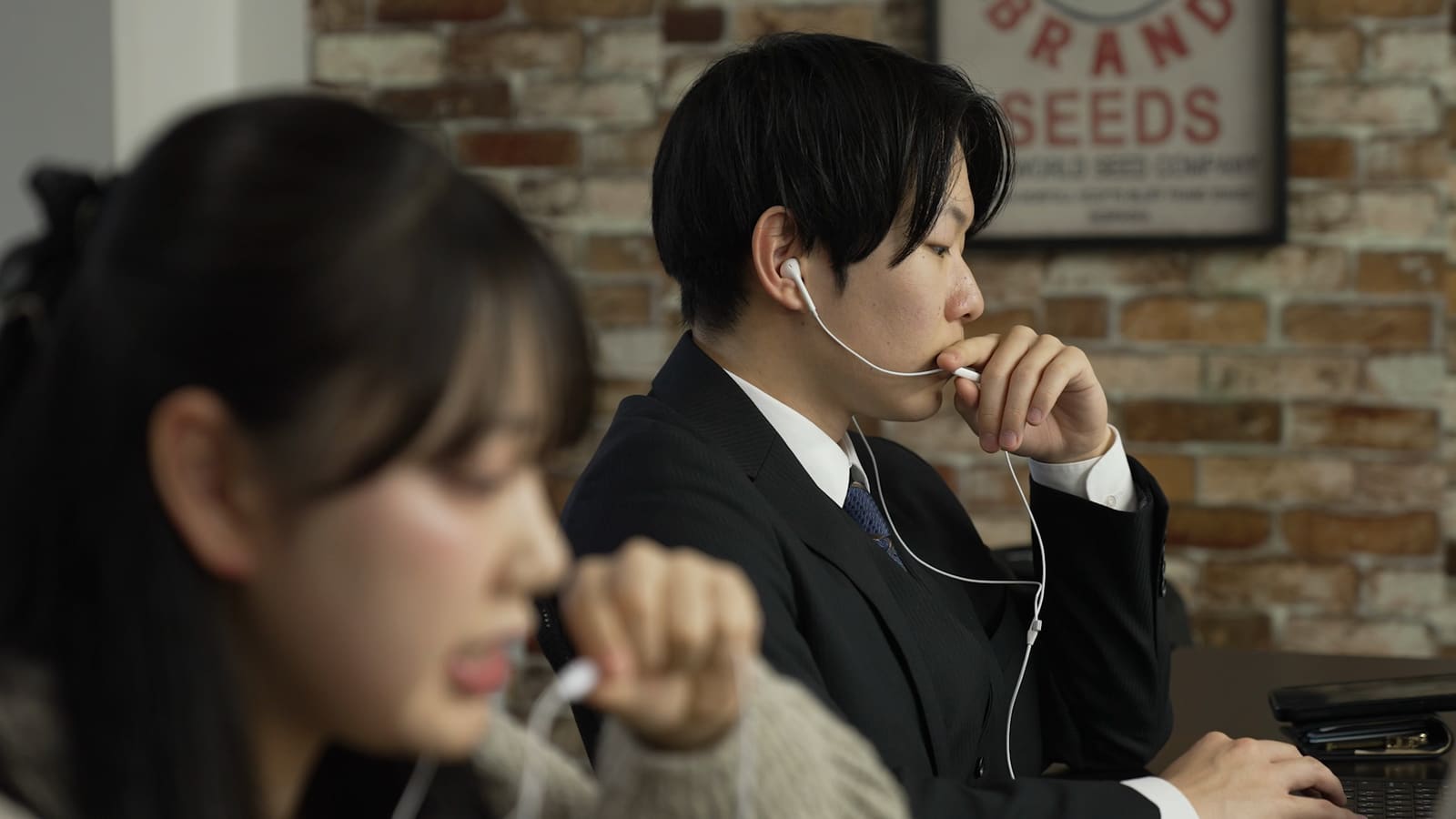 手前で、イヤホンのコードを口元に寄せて話す女性。奥で、イヤホンをつけたスーツの男性が、手を口元に当てて前を見ている