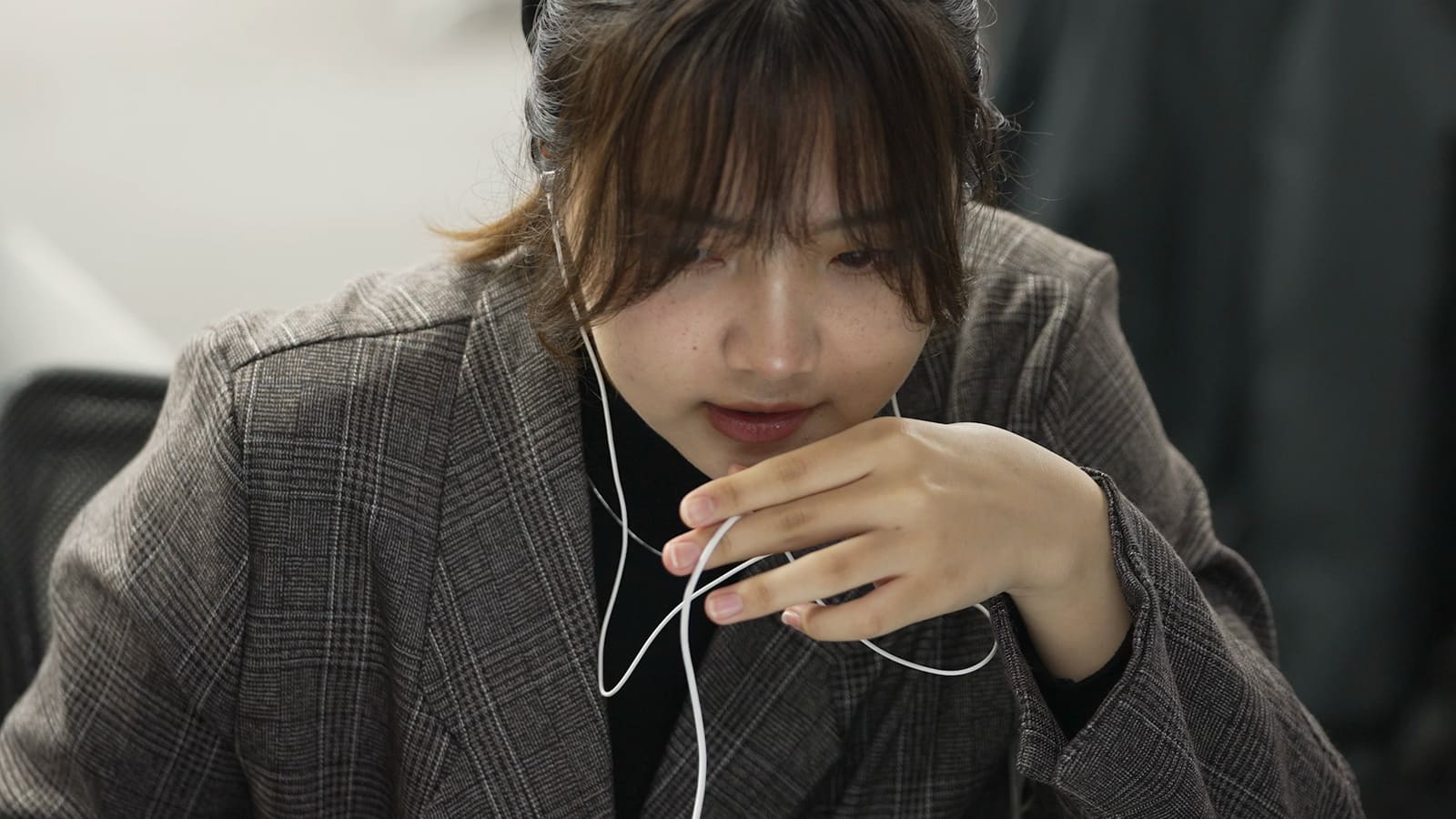 ツイードのジャケットの女性が、イヤホンのコードを片手で口元に寄せ、うつむいて画面を見ている