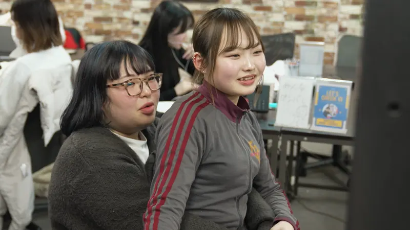 眼鏡の女性と、赤い線の入ったジャージの女性が並んで座り、同じ方向を見ている。ジャージの女性はほほえみ、眼鏡の女性は話している