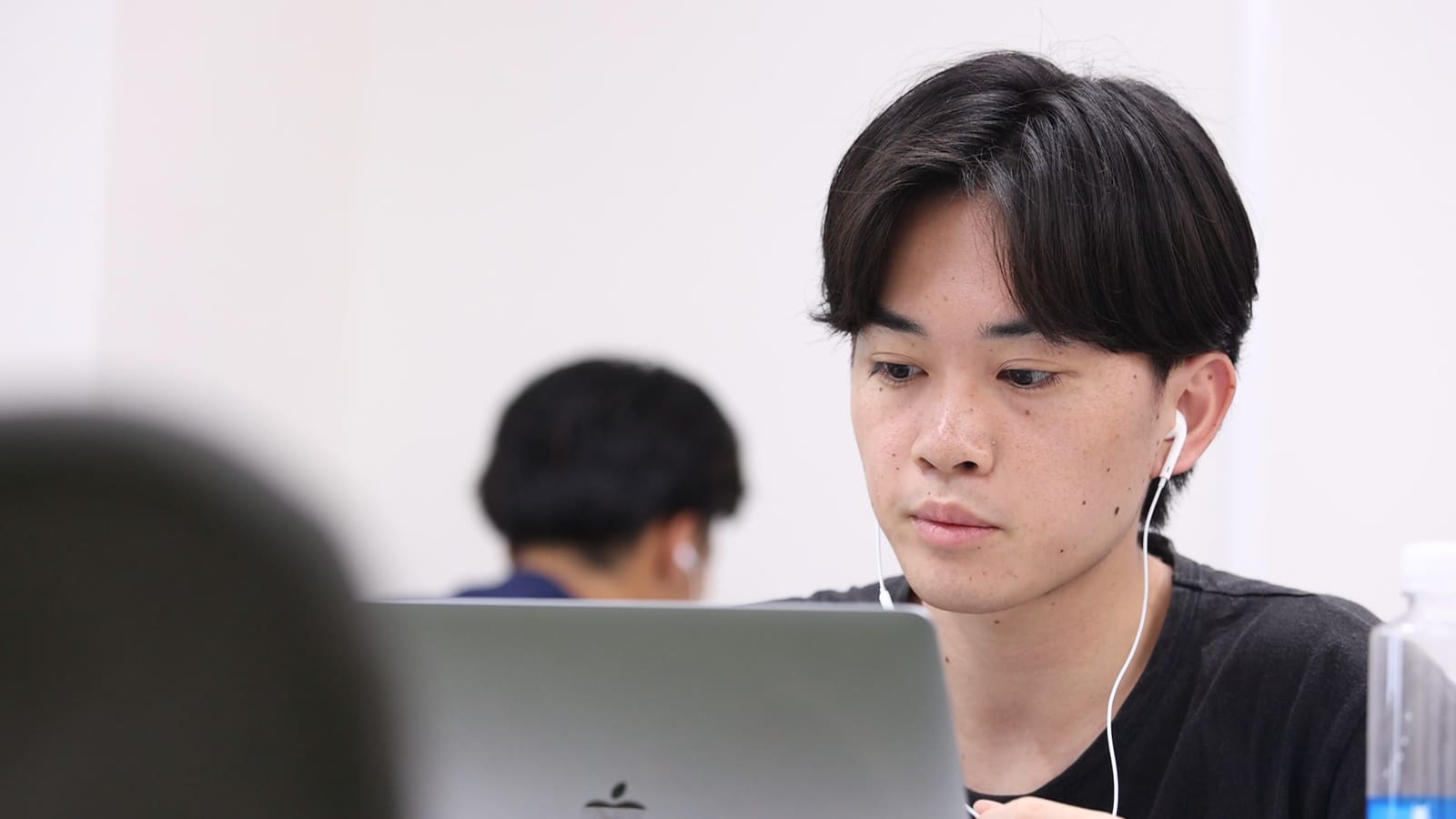 イヤホンをつけた黒いTシャツの男性が、ノートPCの画面を見ている。奥に別のメンバー