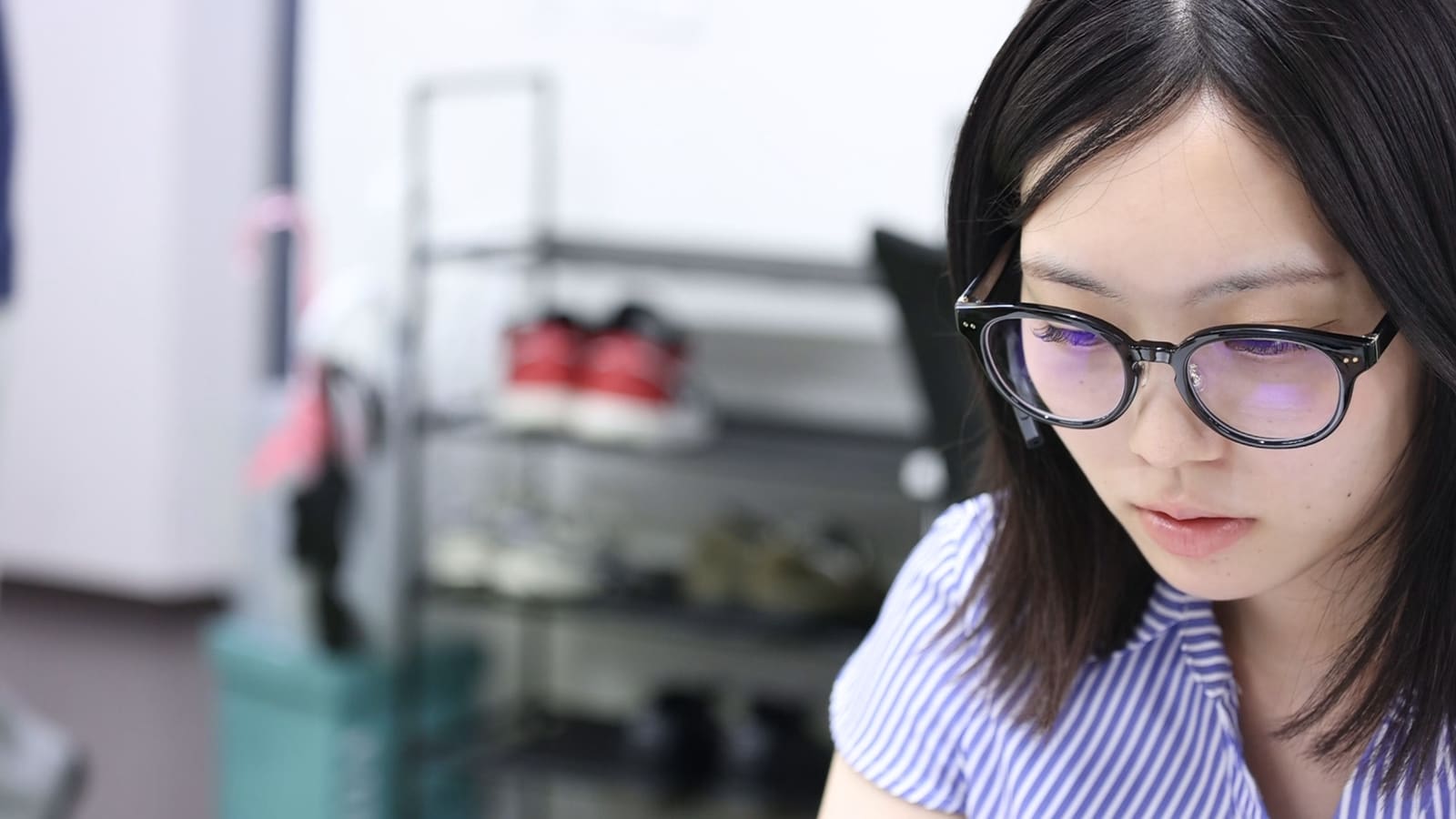 眼鏡をかけた女性が、うつむいて画面を見ている。後ろに靴の棚