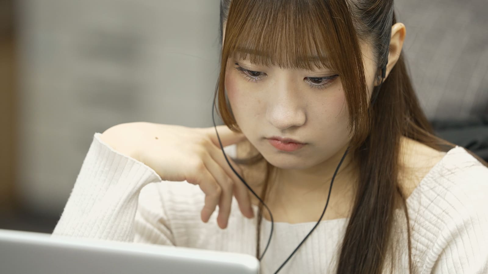 白いニットの女性が、イヤホンをつけて、首元に手を当てて、うつむいて画面を見ている