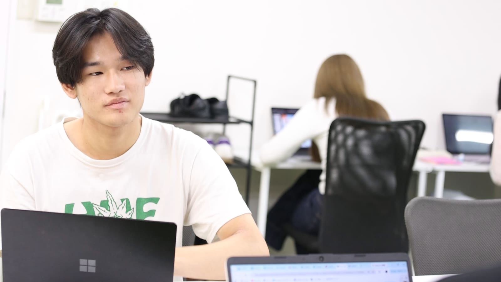 白いTシャツの男性が、ノートPCの前に座り、顔を上げて横のほうを見ている。奥の席に女性