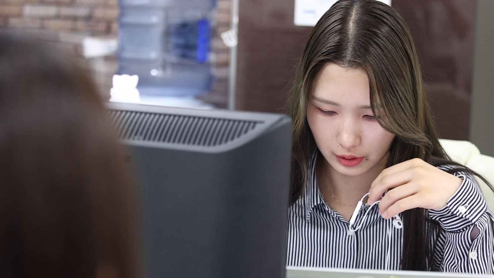 ストライプのシャツの長い髪の女性が、イヤホンのコードを指でつまんで、うつむいて画面を見ている