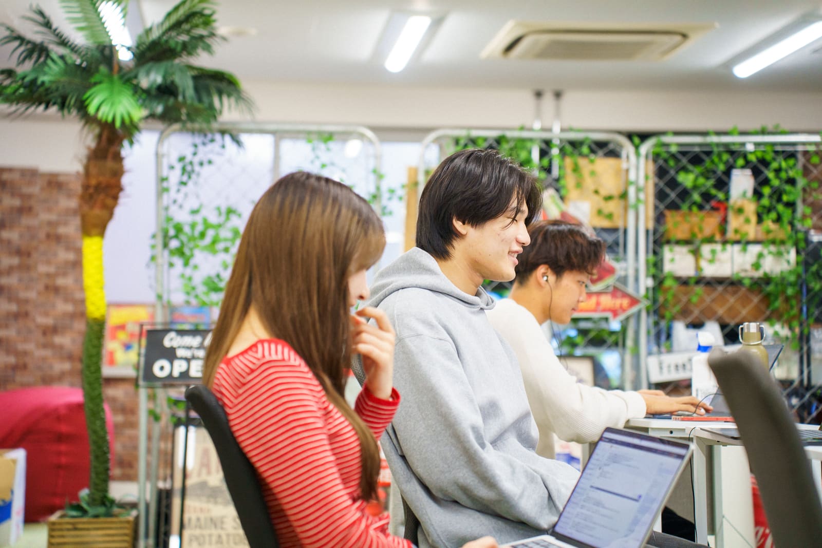 ヤシの木の飾りのそばで、3人がそれぞれ作業している。赤いボーダーの女性は頬に手を当てて画面を見ている