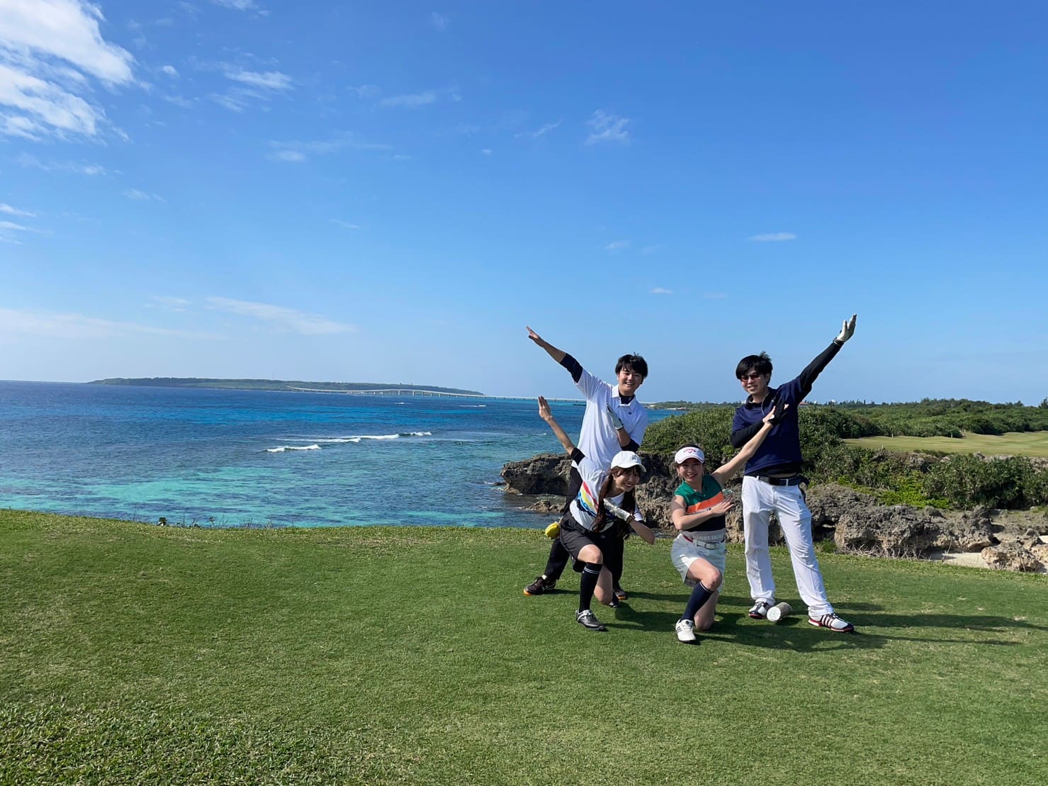 海沿いのゴルフ場の芝生で、数人がそれぞれ両腕を広げてポーズをとっている