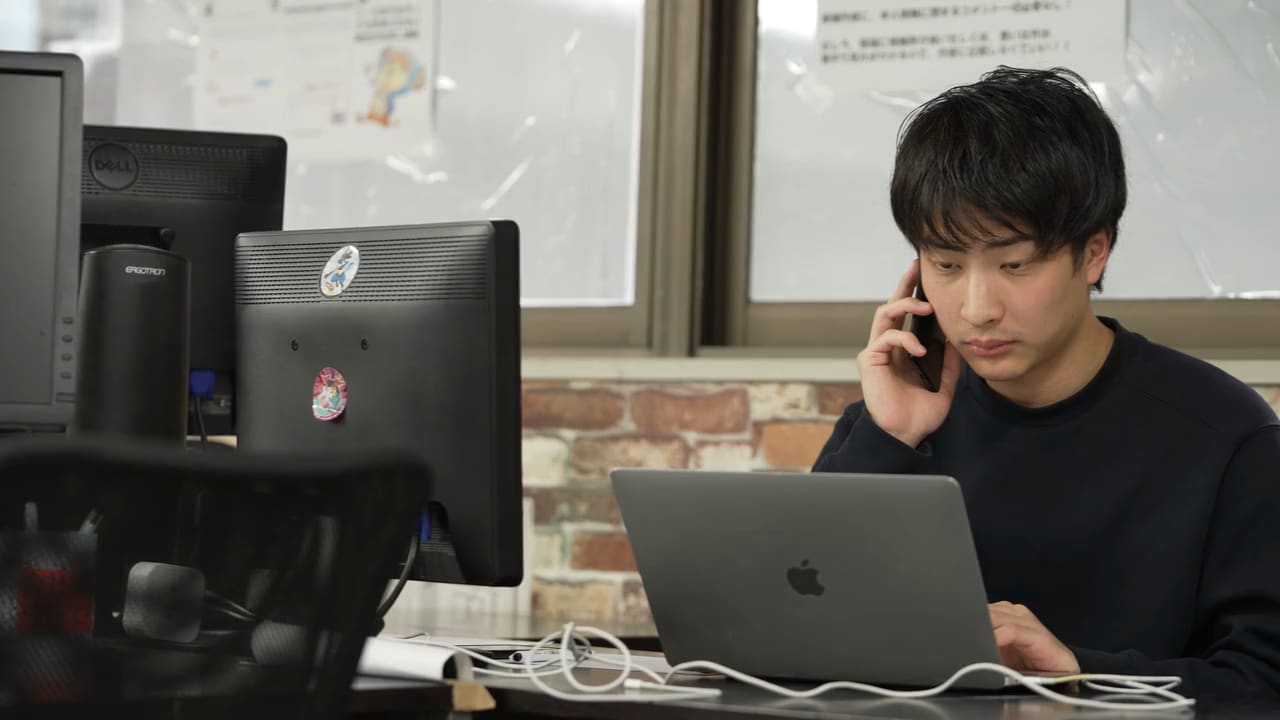 紺のスウェットの男性が、窓とレンガの壁を背にして、スマホを耳に当てて話しながら、ノートPCを操作している