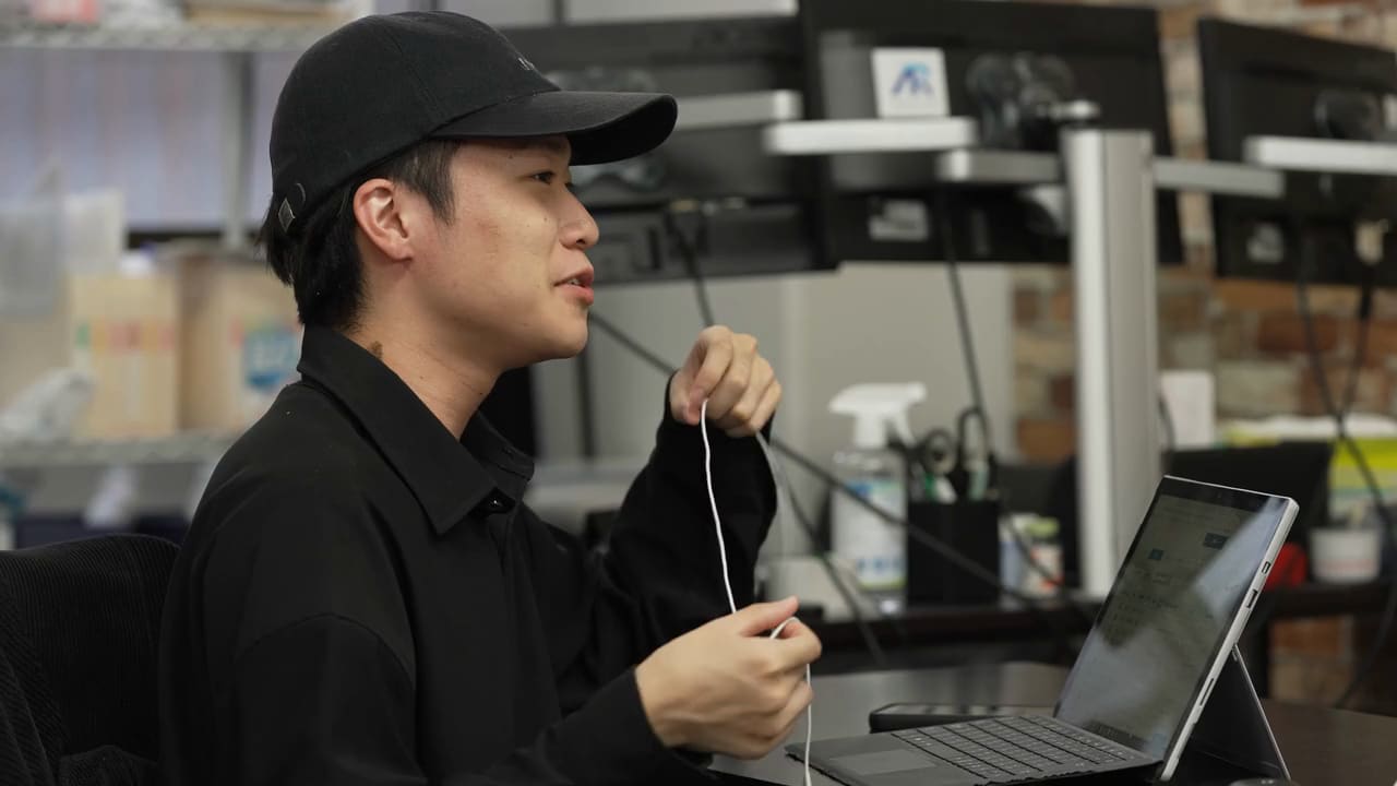 黒いキャップに黒いシャツの男性が、イヤホンのコードを両手に持って、笑いながら話している